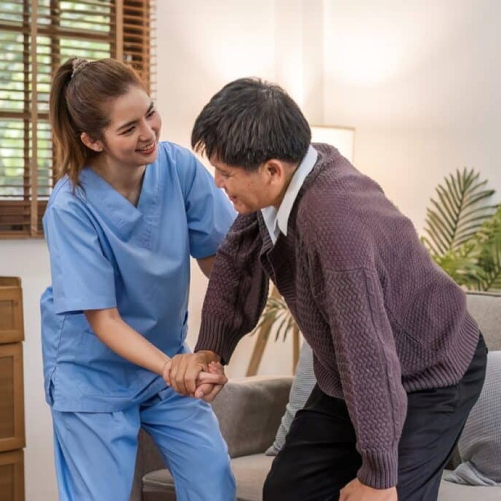 A caring young Asian nurse helps an elderly person walk in the house. An elderly disabled man tries to walk with the help of a female caregiver..
